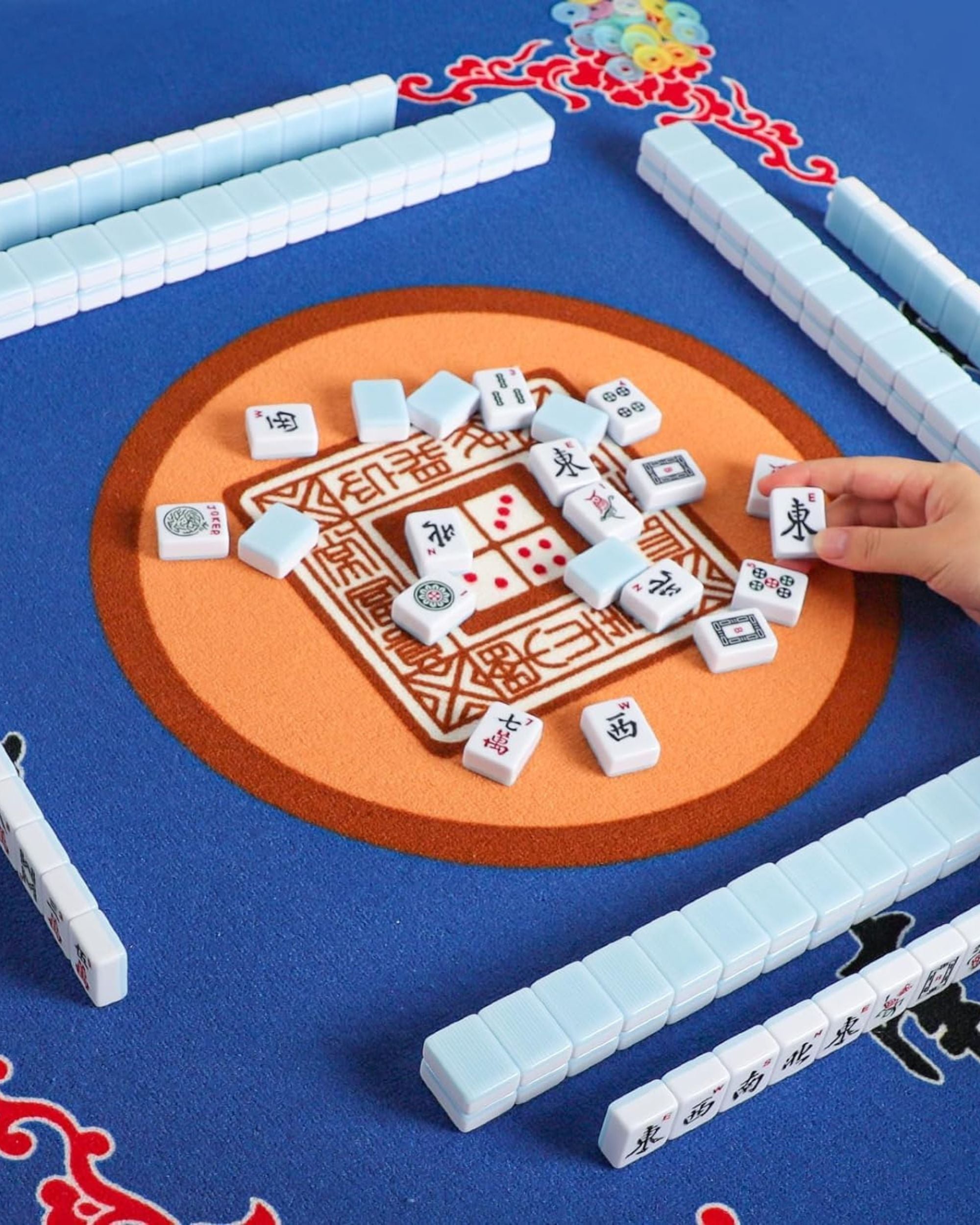 Mahjong tiles on a blue mat with tile racks and a hand selecting a tile