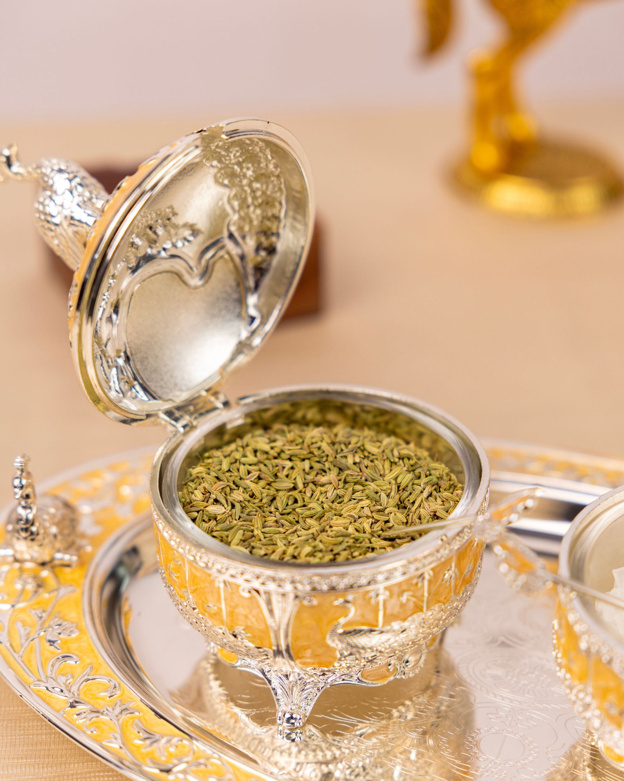 Open silver-and-gold serving bowl with peacock lid ornament, filled with green dried herbs