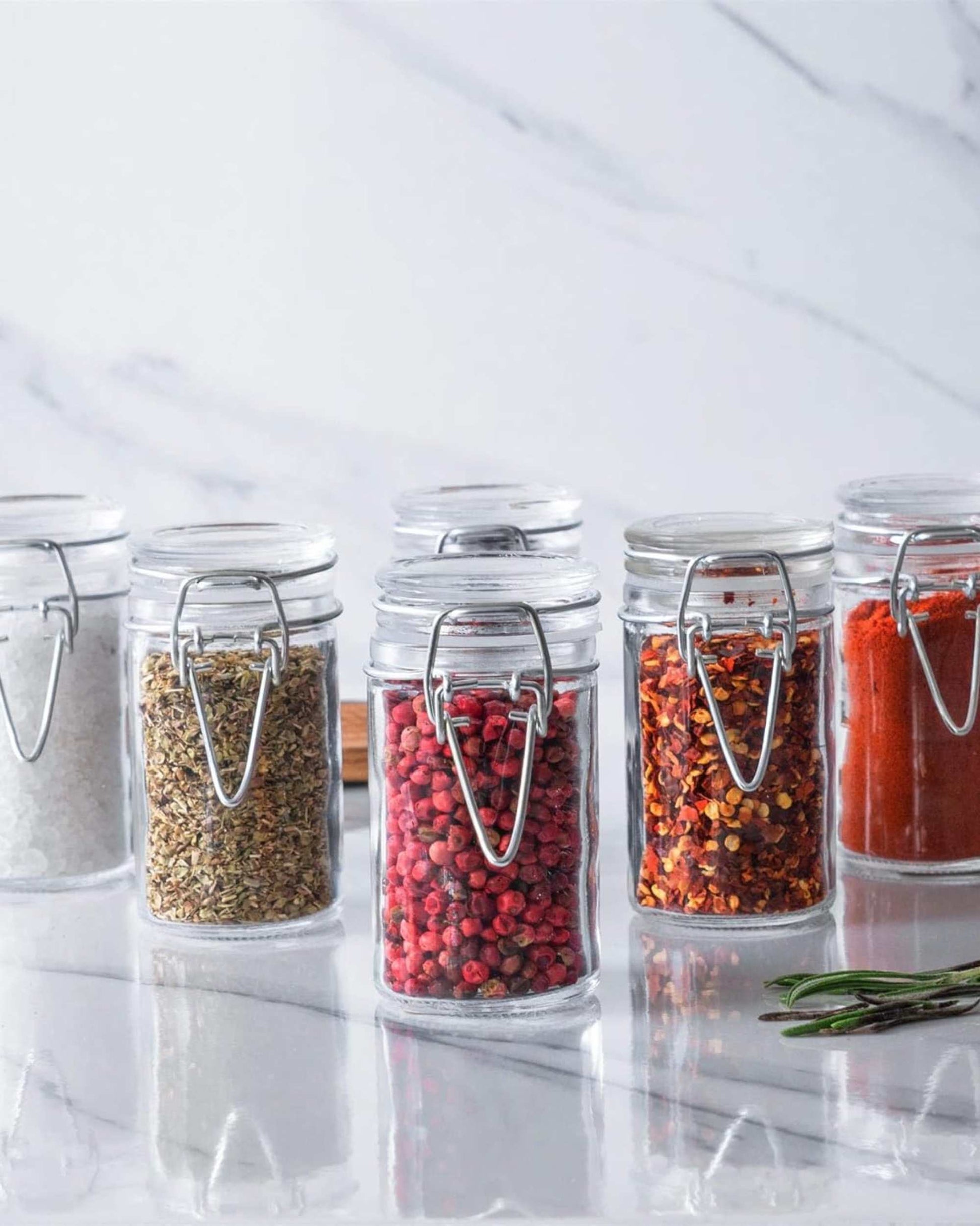 Row of clear glass clip-lock jars filled with assorted spices and seeds