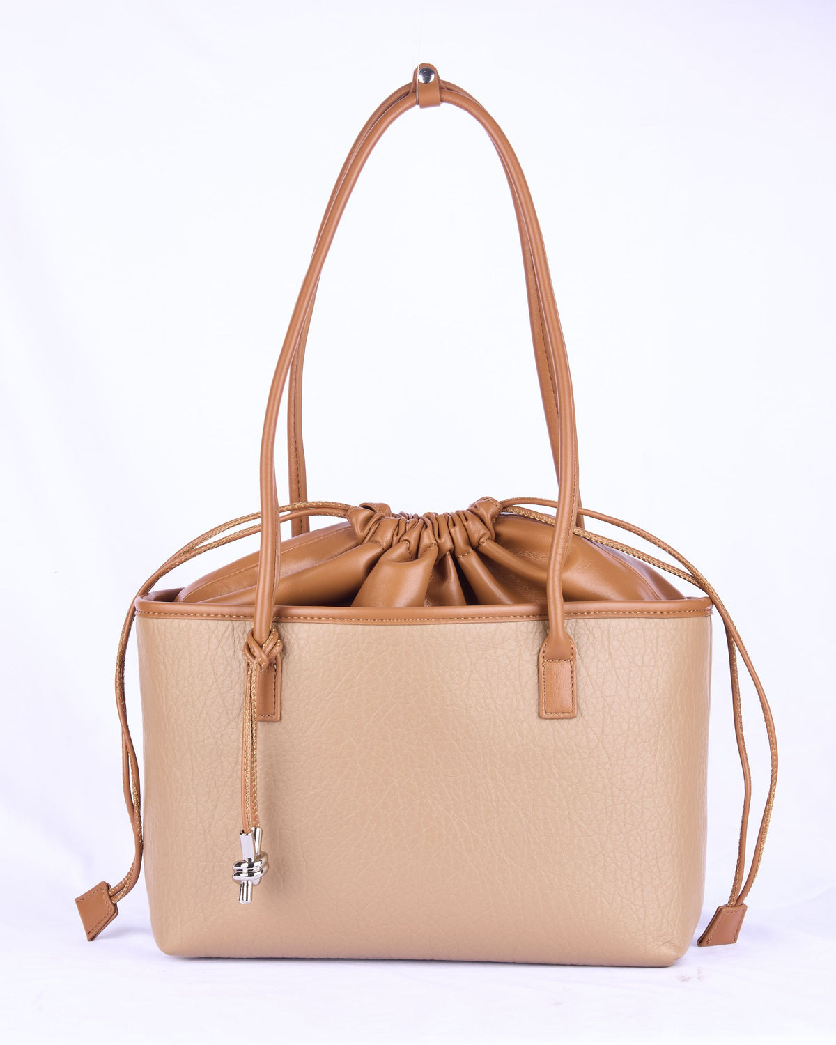 A structured tote bag in beige pebbled leather with tan leather handles, a drawstring closure, and a silver cross charm, shown on a white background.