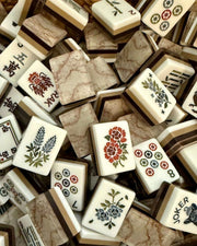 A pile of Mahjong tiles with cream-colored faces, brown marble-patterned backs, and colorful traditional Chinese illustrations including floral designs and symbols.