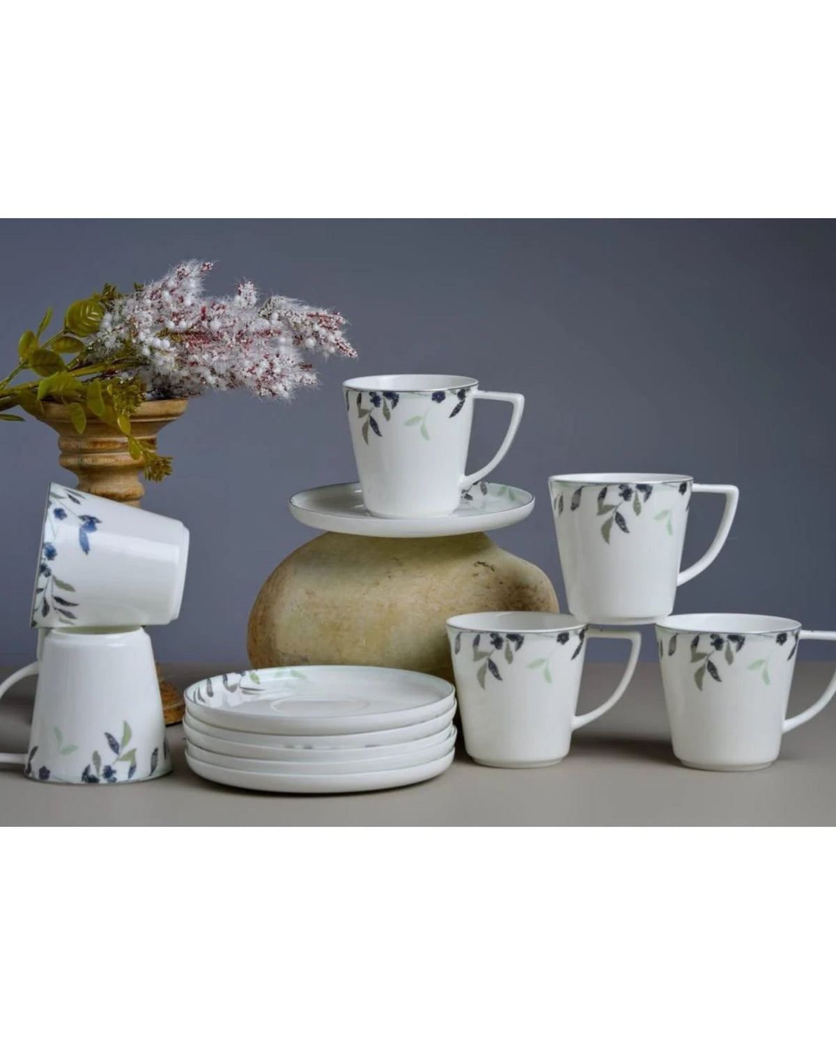 White coffee mugs with blue and green floral print beside matching plates and saucers.