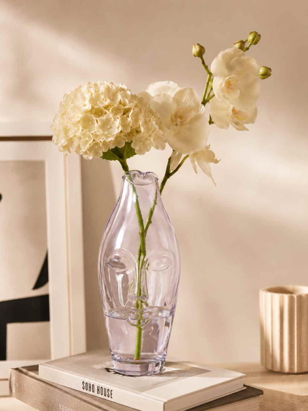 Clear glass face vase with cream-white flowers and green stems on a book