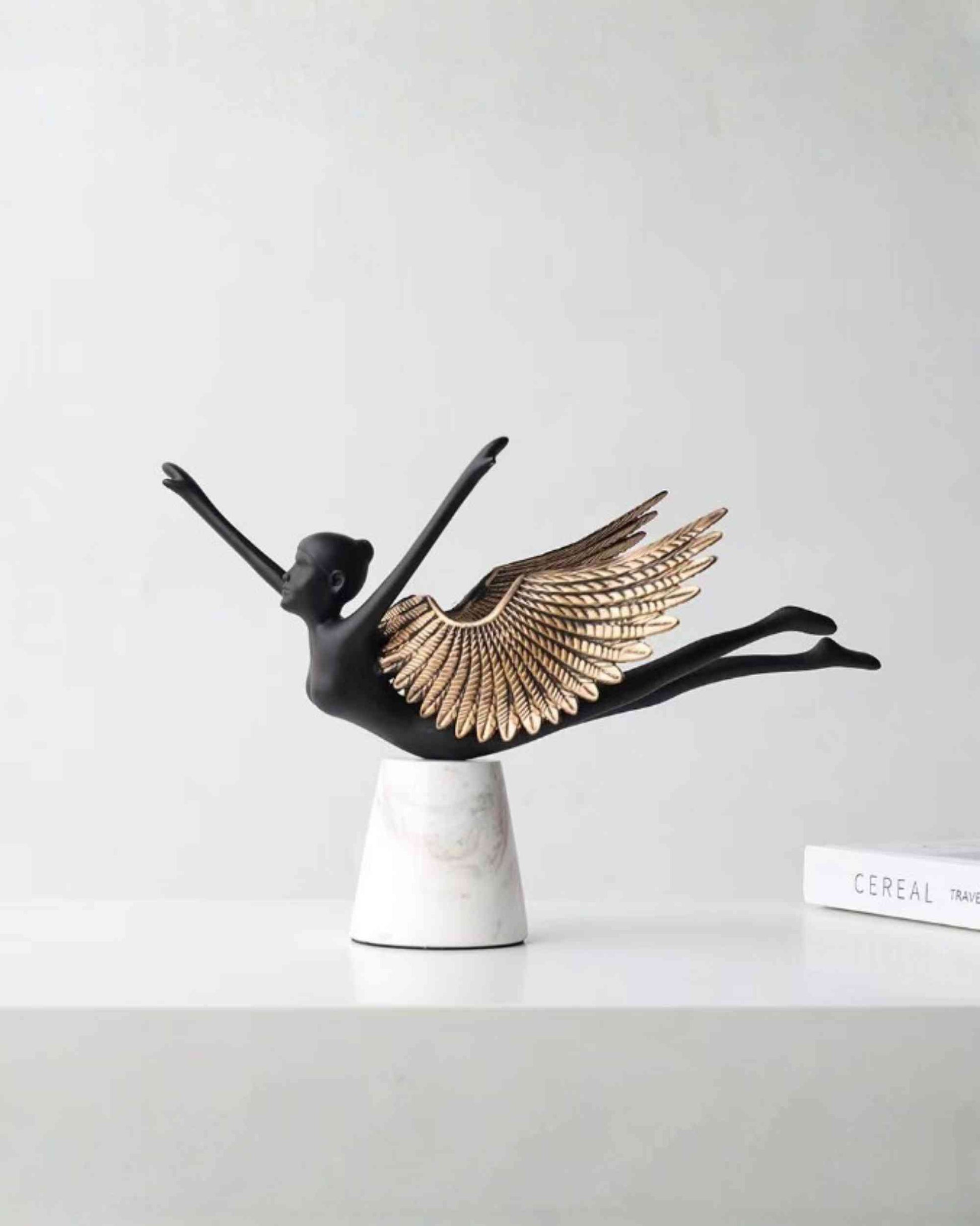 Winged bird form sculpture with black metal body, gold feather wings, on white pedestal base